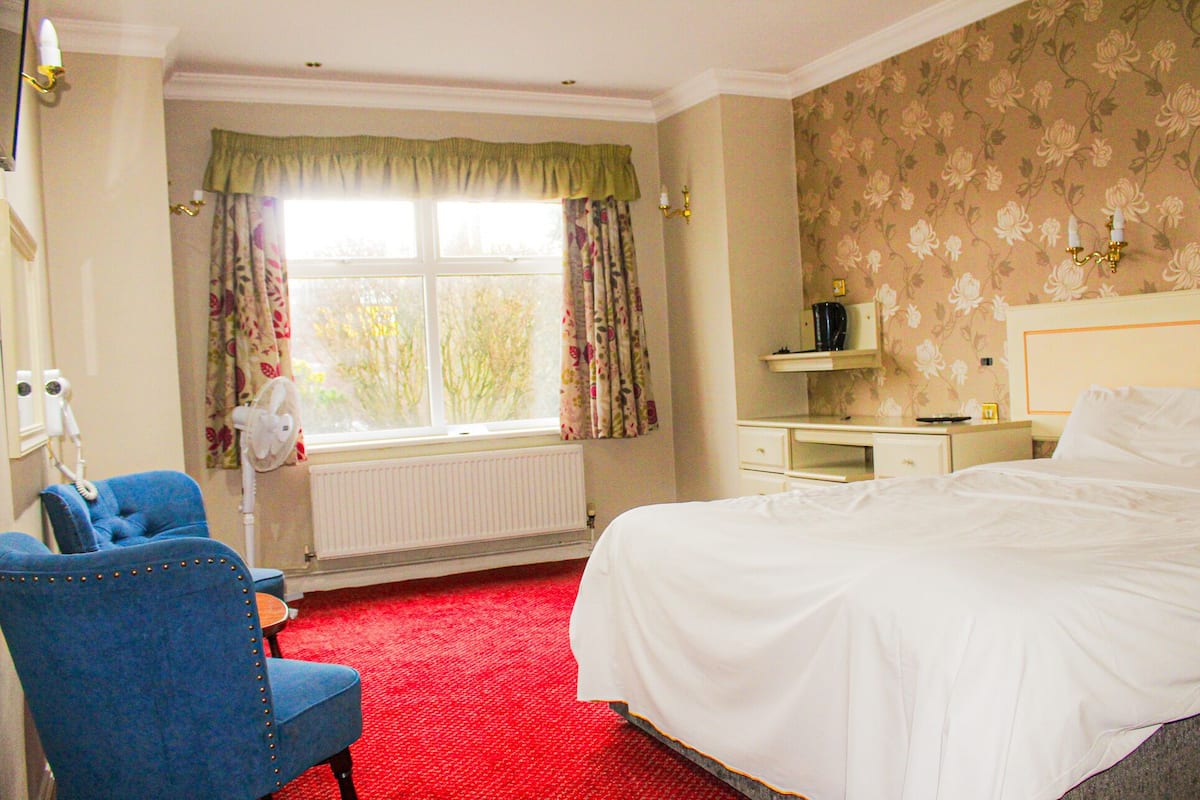 A hotel room with a large bed, a desk with a chair, a radiator, a window with curtains, and a wall with floral wallpaper.