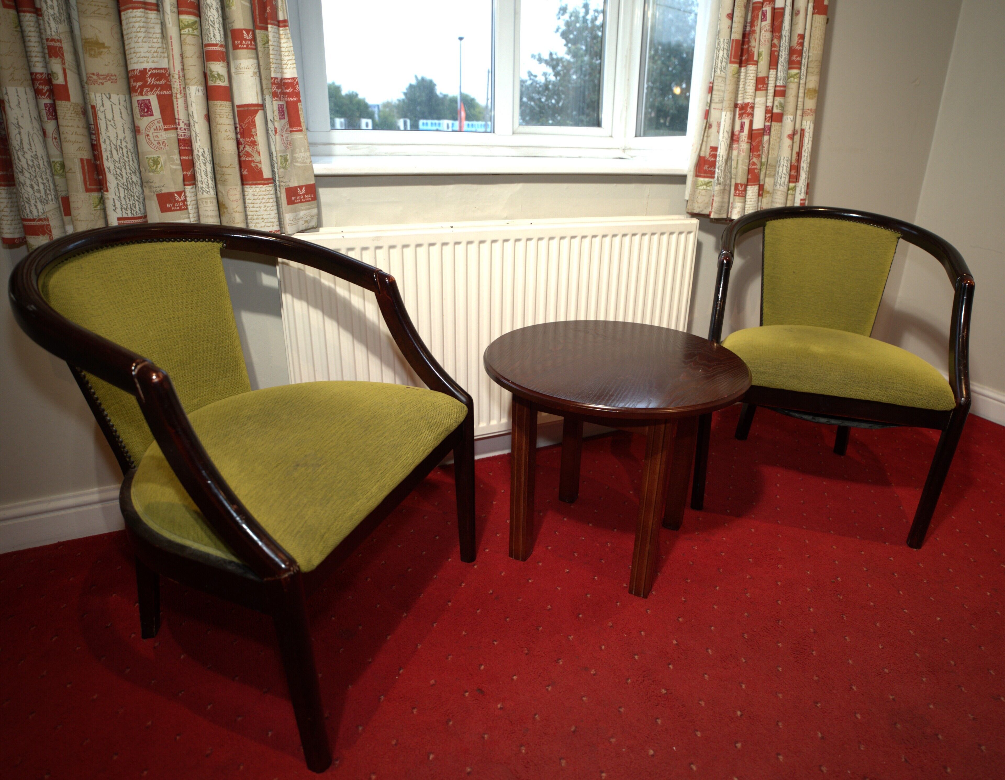 Two green upholstered chairs with wooden frames and a small round wooden table in a room with a red carpet and a window with curtains.