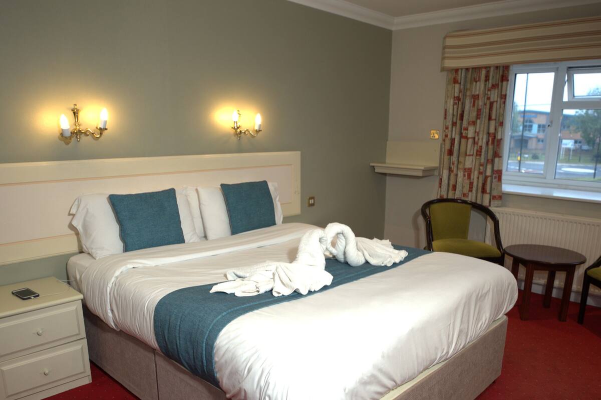 A neatly made bed with a blue blanket and white pillows, a bedside table with a phone, and a chair by a window with curtains.