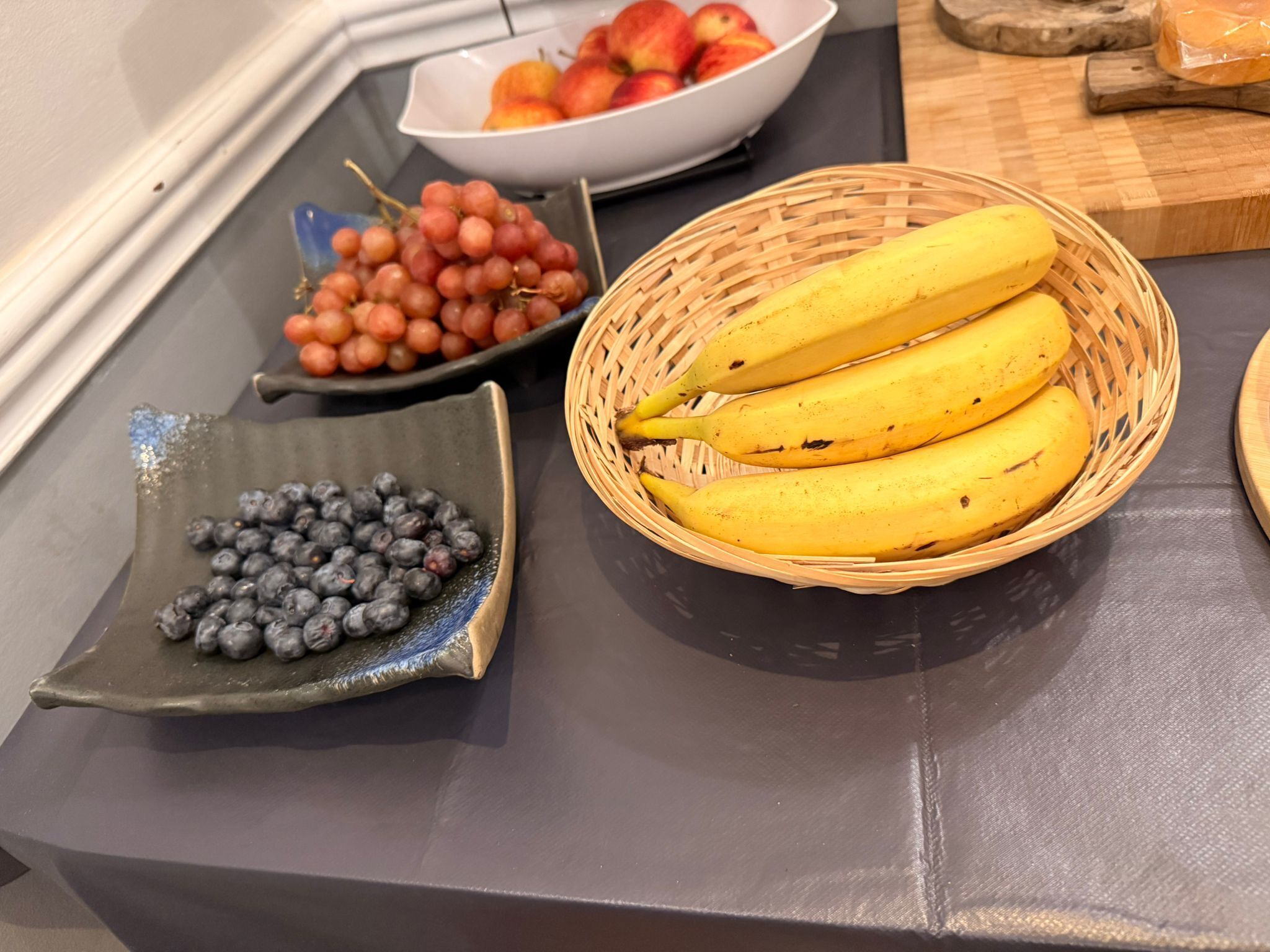 A table with a bowl of grapes, a bowl of peaches, a plate of blueberries, and a basket of bananas.