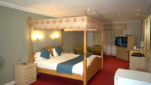 A four-poster canopy bed with a wooden frame and a patterned canopy, a bedside table with a lamp, a green chair, a television, and a dresser. - Consort Hotel & Urban Spa (Rotherham)