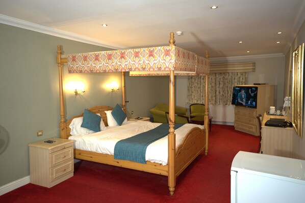 A four-poster canopy bed with a wooden frame and a patterned canopy, a bedside table with a lamp, a green chair, a television, and a dresser. - Consort Hotel & Urban Spa (Rotherham)