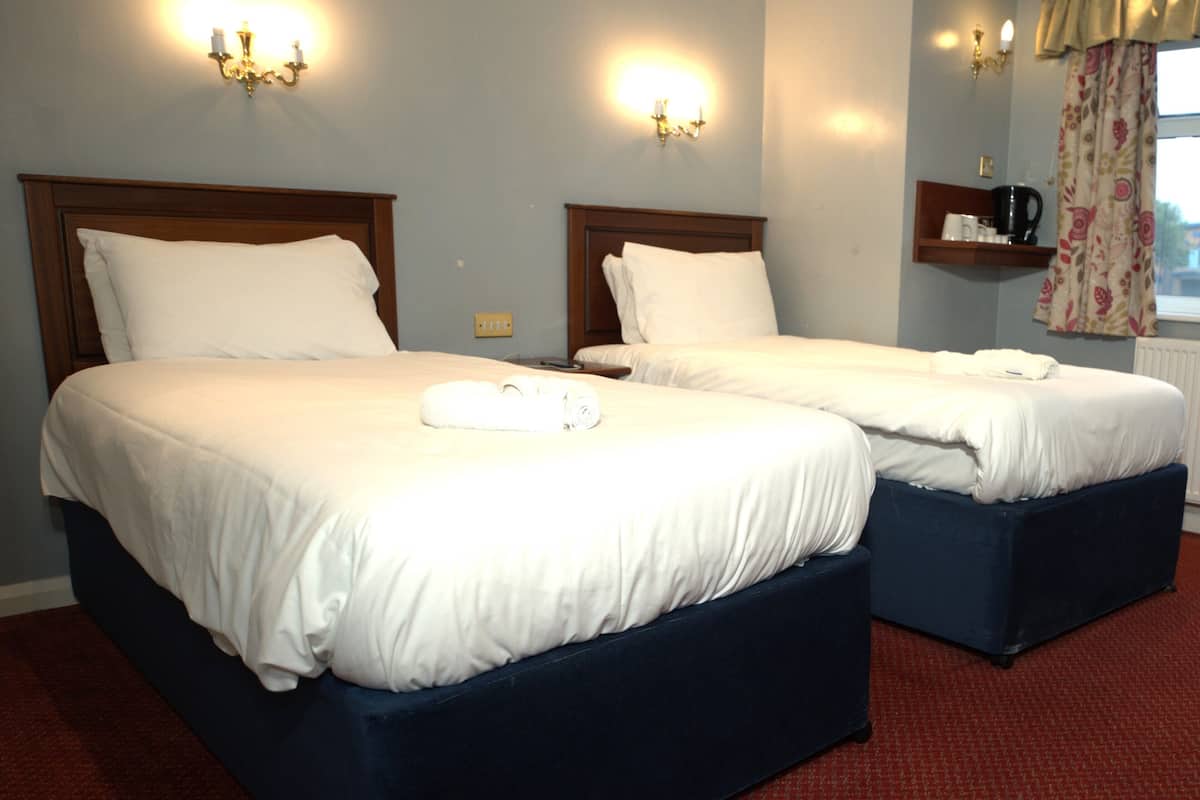 A hotel room with two beds, each with white linens and a rolled towel on the bed. There are wall sconces with lights, a window with curtains, and a shelf with a coffee pot.