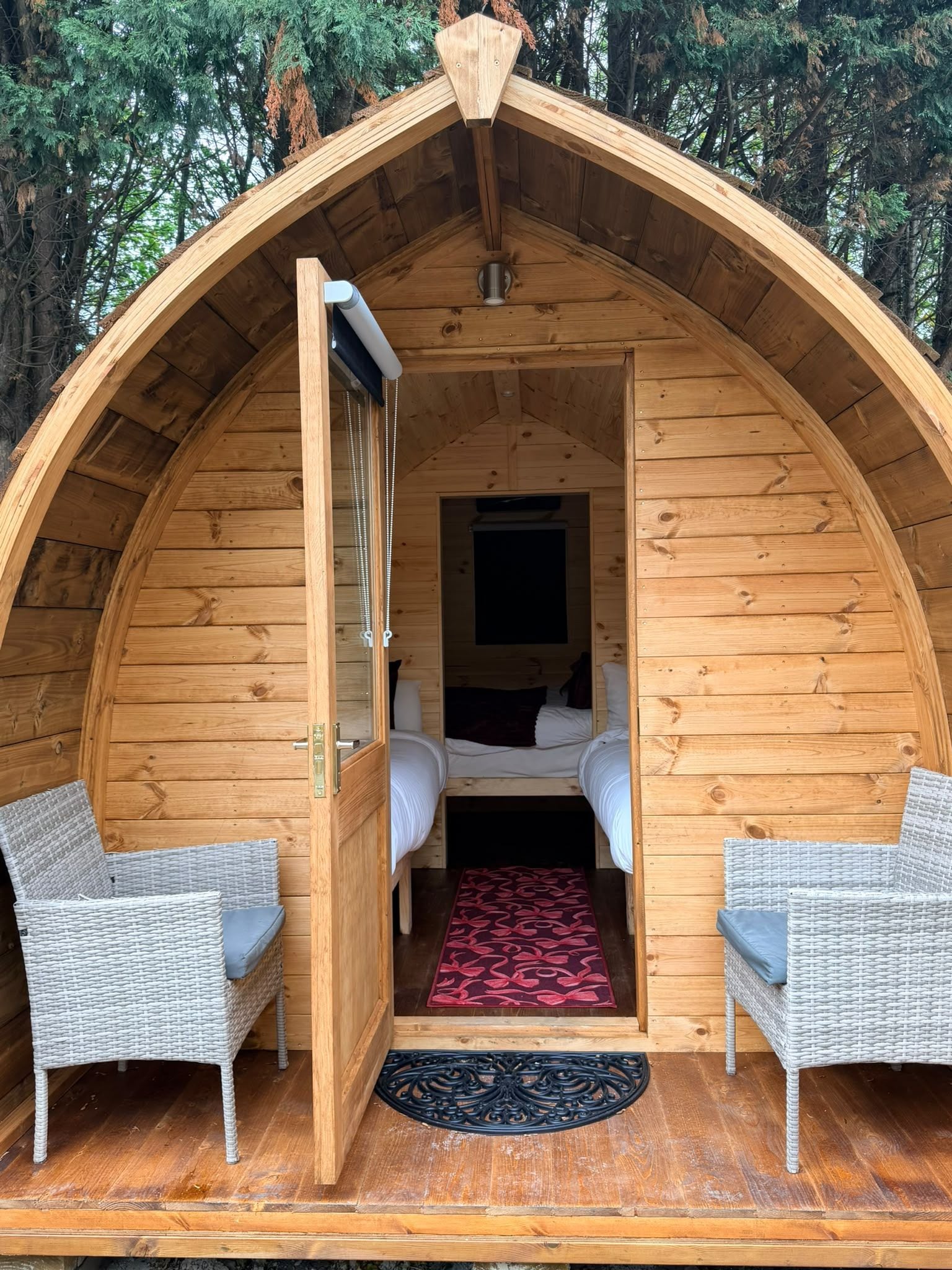 Una cabaña de madera acogedora con techo curvo, una cama, dos sillas de mimbre y un tapete rojo estampado.
