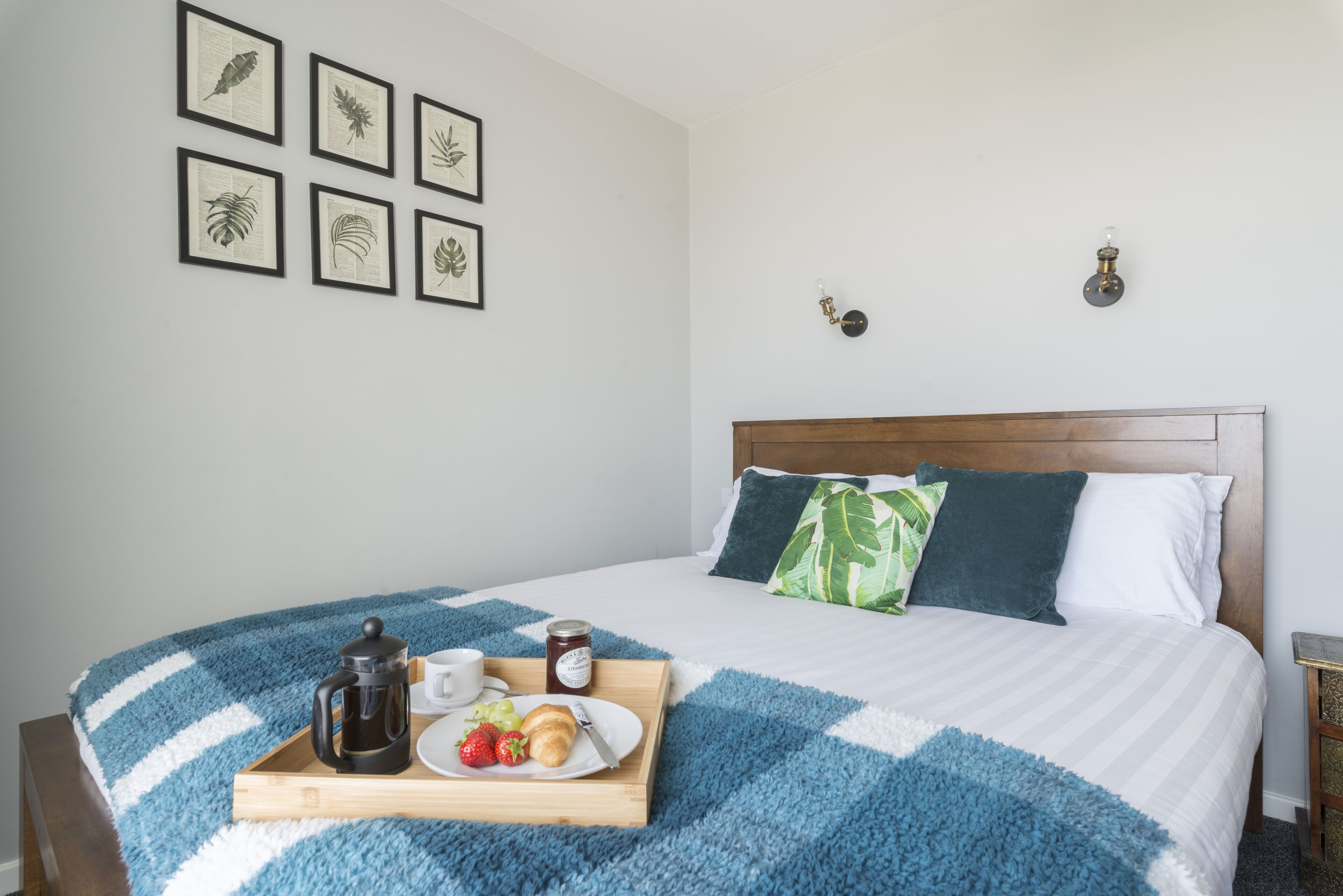A neatly made bed with a blue and white striped blanket, a wooden bedside table with a coffee pot, a tray with food, and framed botanical prints on the wall.