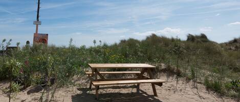 A wooden picnic table in a sandy area with wildflowers and grasses.