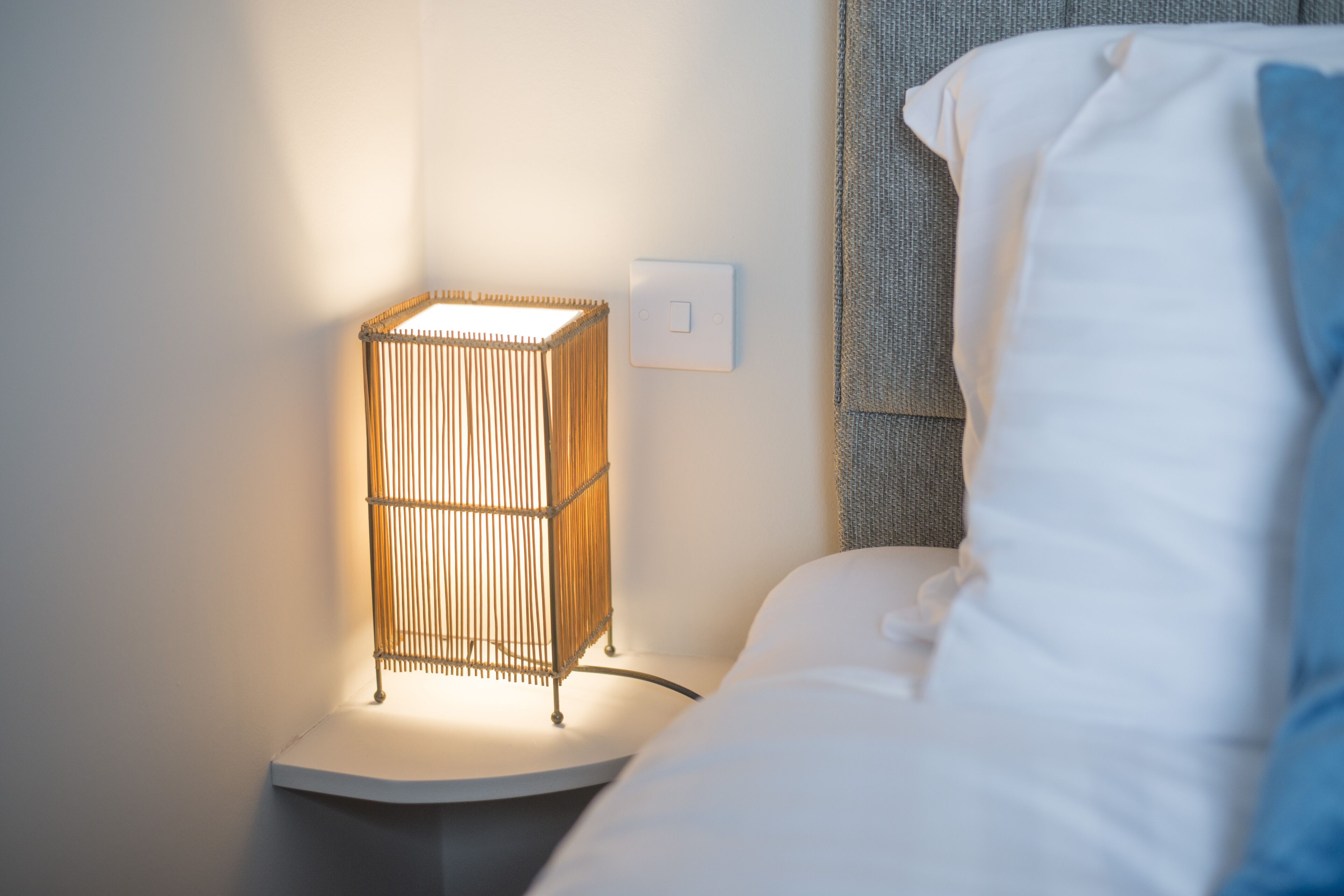 A bedside table with a lamp, a power outlet, and a bed with white bedding.