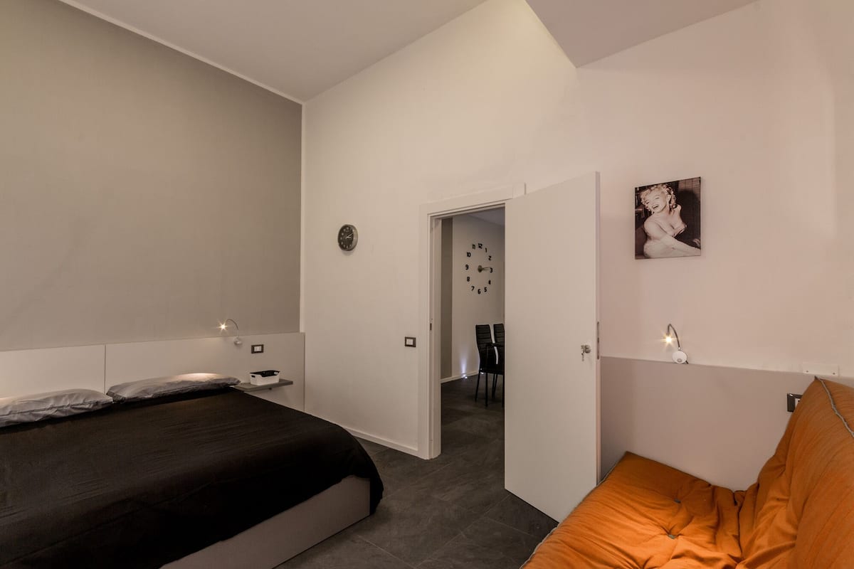 Un dormitorio con una cama, una silla, una puerta que da a otra habitación y un reloj de pared.