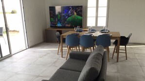A modern living room with a grey sofa, a wooden dining table with blue chairs, and a large flat-screen TV on a stand. - House the air of the wind (Saint-Denis-d'Oléron)