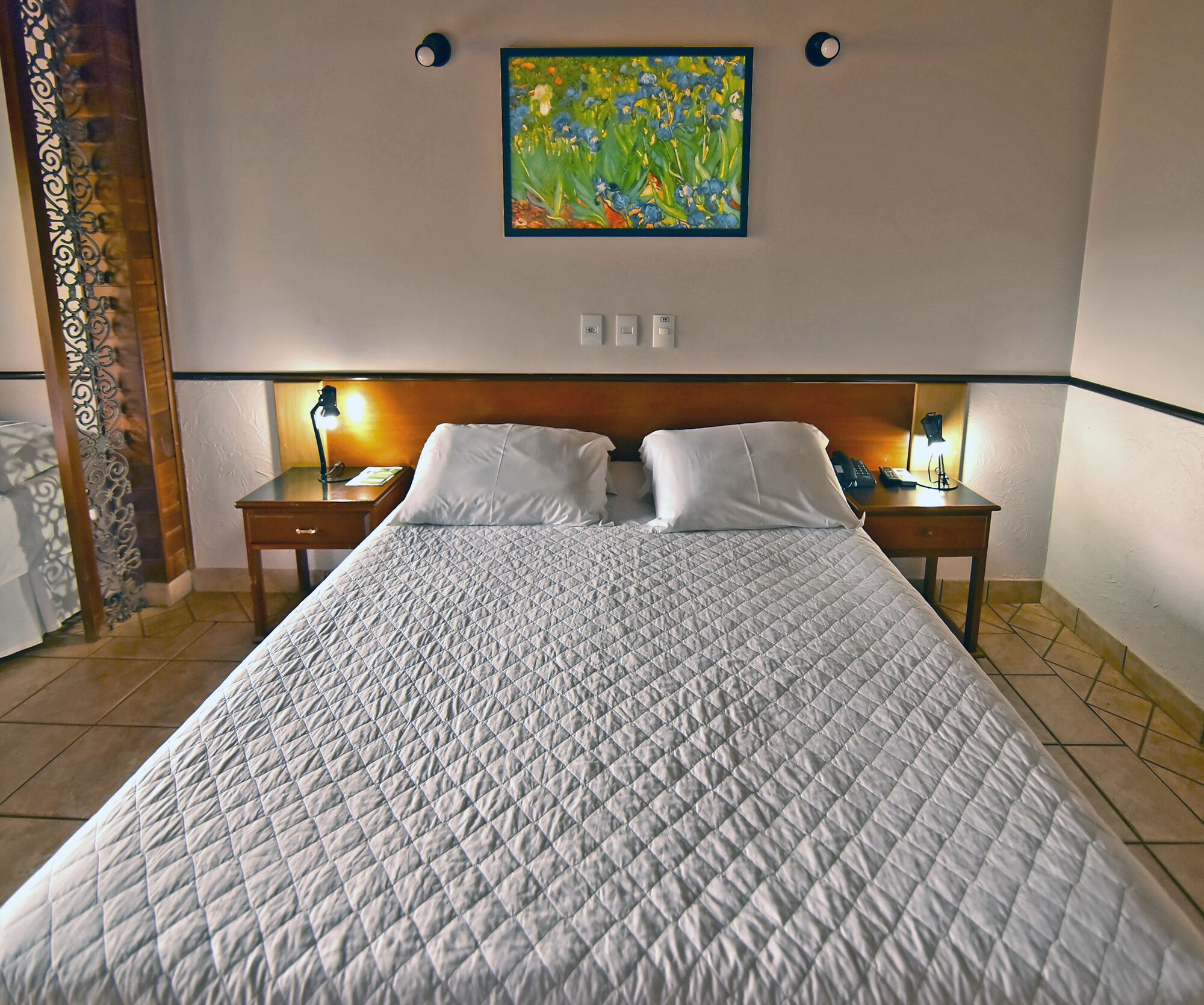 Una cama bien hecha con cubrecolchón acolchado, dos mesitas de noche con lámparas, un cuadro en la pared y una cortina decorativa.