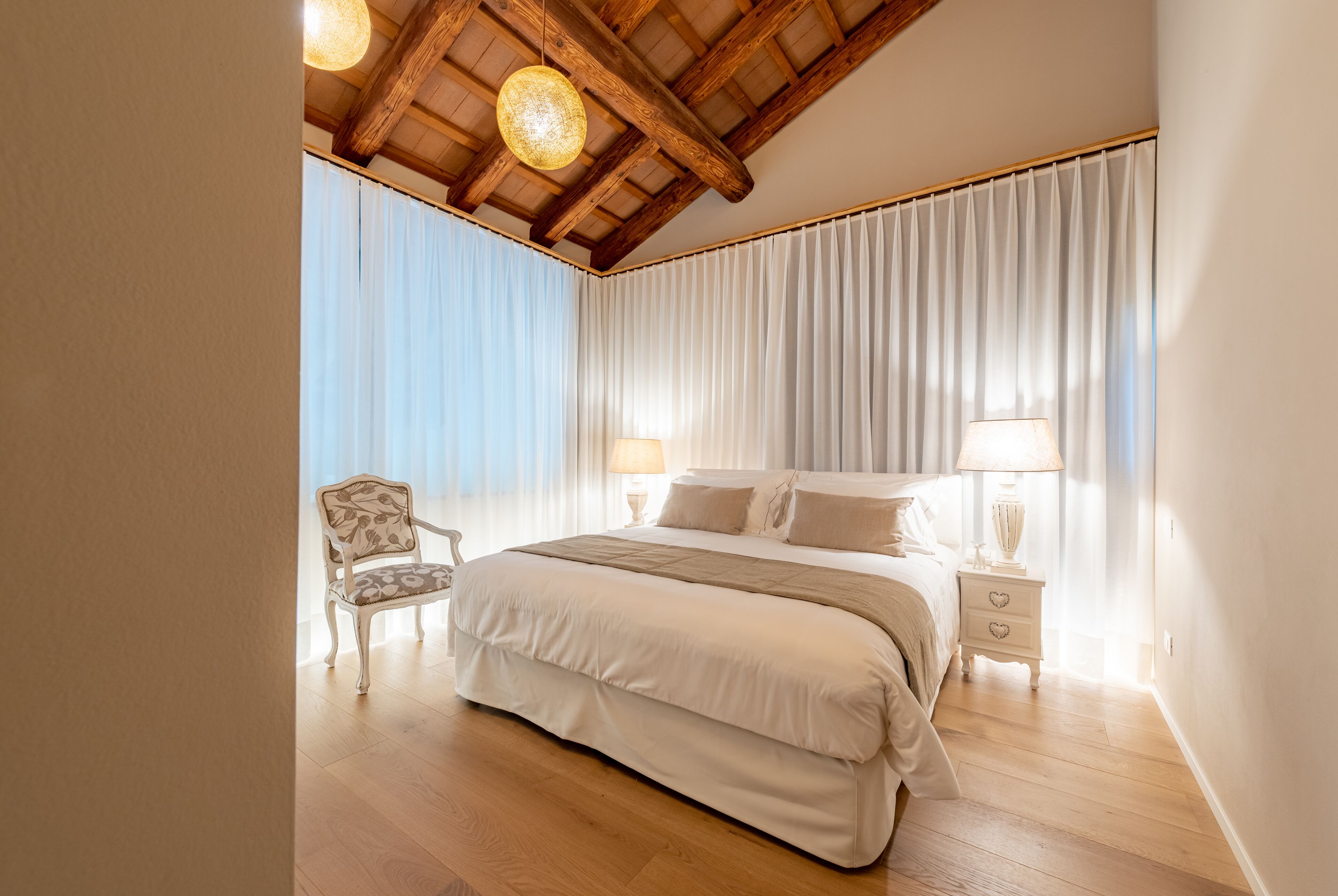 A bedroom with a large bed, wooden beams on the ceiling, a chair, and a nightstand with a lamp.
