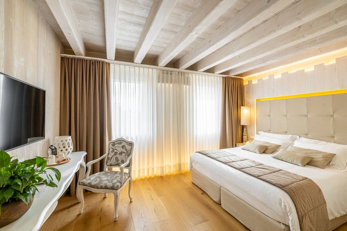 A hotel room with a bed, a desk with a TV, a chair, and a large window with curtains.