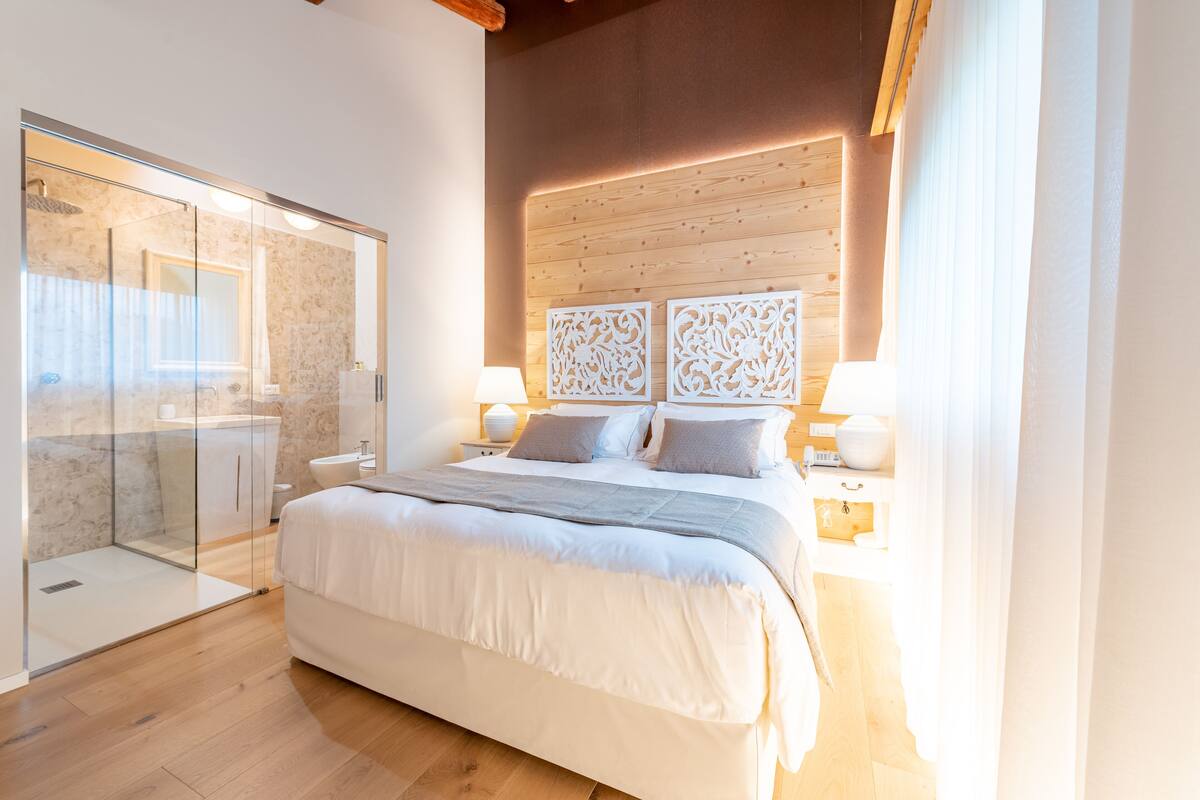 A modern bedroom with a large bed, bedside tables, a glass shower enclosure, and a wooden ceiling.