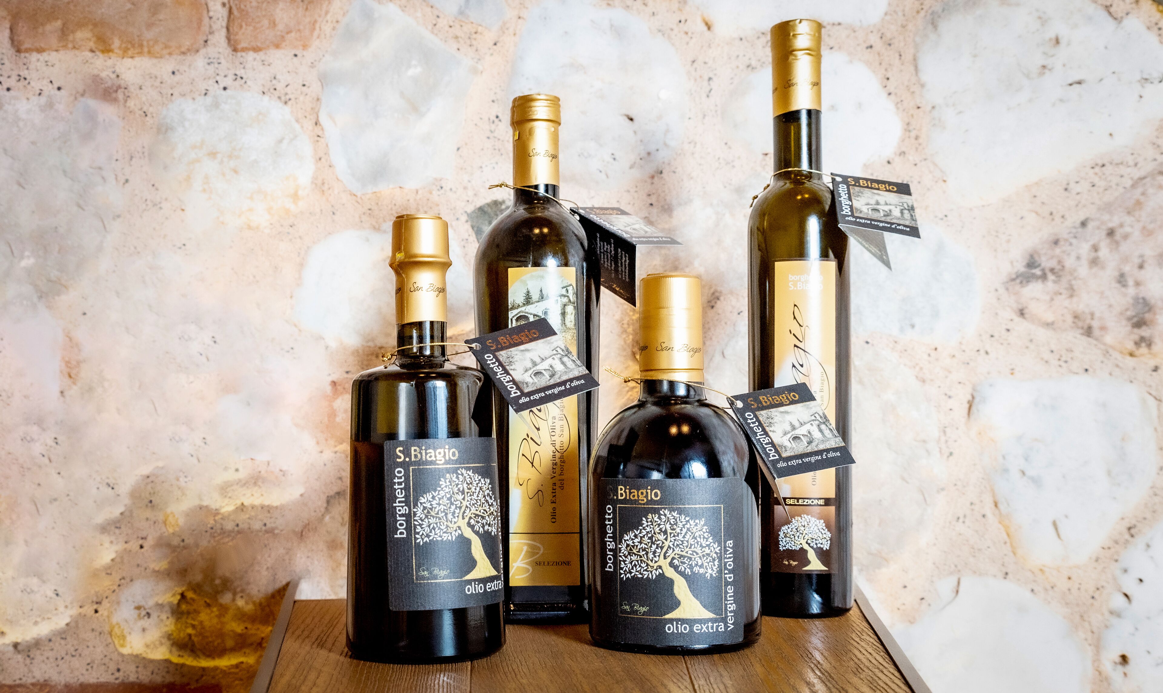 Cuatro botellas de aceite de oliva virgen extra S. Biagio sobre una superficie de madera frente a una pared de piedra.