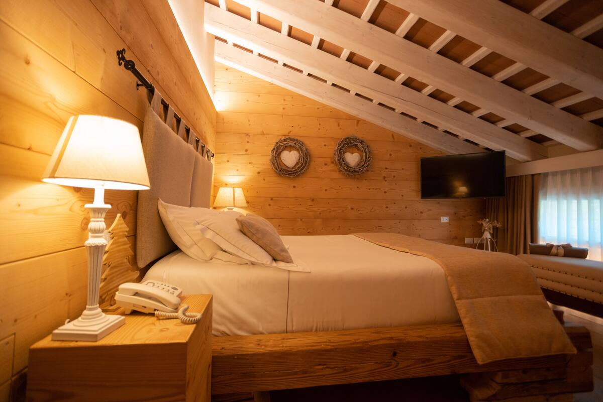 A cozy bedroom with a wooden ceiling, a bed with white linens, a bedside lamp, a telephone, and a flat-screen TV mounted on the wall.