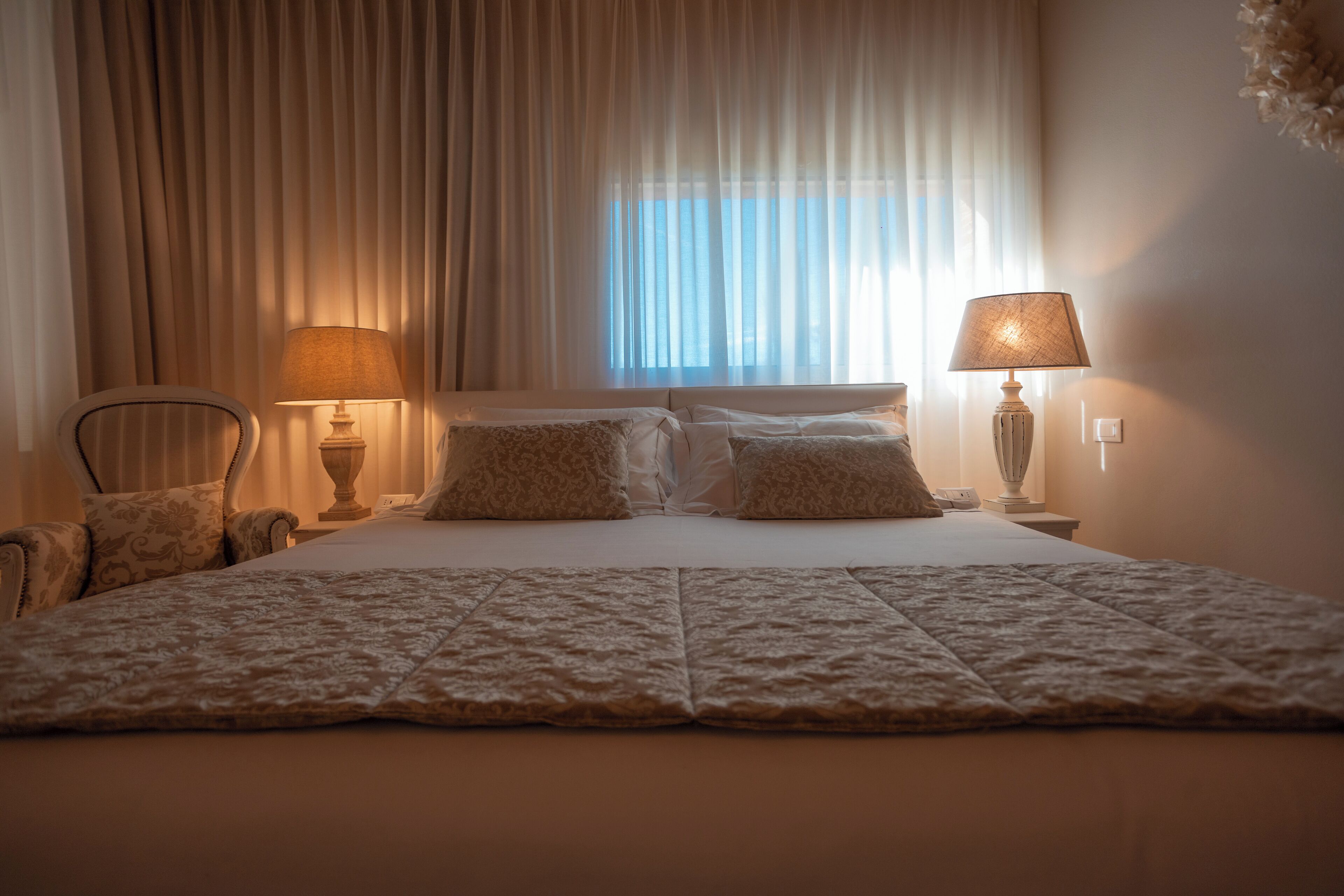 Ein ordentlich bezogenes Bett mit einem gemusterten Überwurf, zwei Nachttischlampen, ein Sessel und ein Fenster mit durchsichtigen Vorhängen.