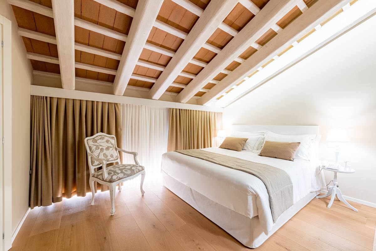 A bedroom with a wooden ceiling, a bed with white bedding, a chair, and a bedside table.