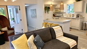 A modern living room with a grey sofa, yellow and patterned cushions, a wicker chair, and a kitchen area with white cabinets, a microwave, and pendant lights. - South Oceanside 1br/1bath, clean and beach close (Oceanside)