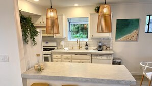 A modern kitchen with white cabinets, a central island with stools, pendant lights, and a window with a view of greenery. - South Oceanside 1br/1bath, clean and beach close (Oceanside)