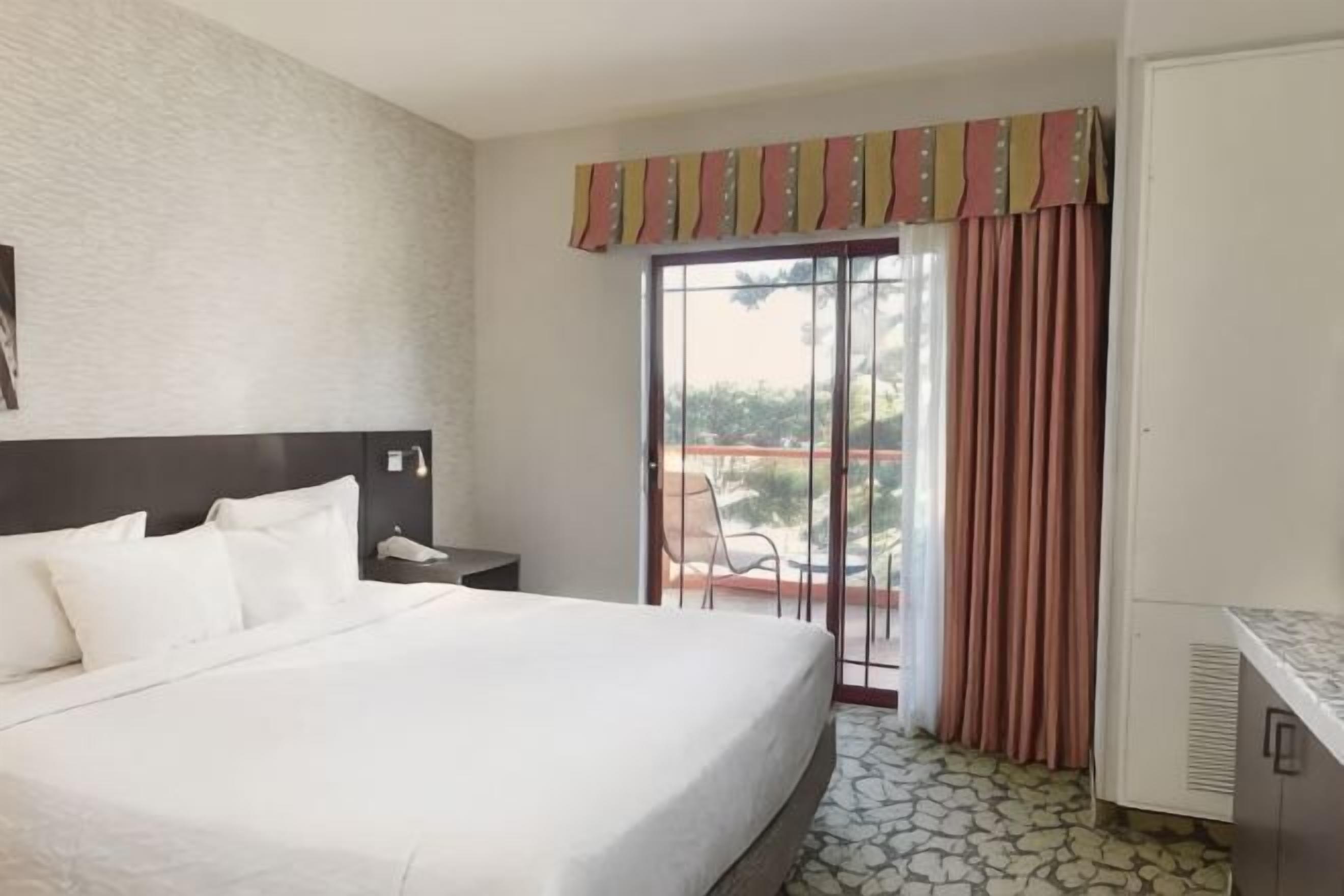 Une chambre d’hôtel avec un grand lit, un bureau, une chaise, une fenêtre avec des rideaux et une vue sur l’extérieur.