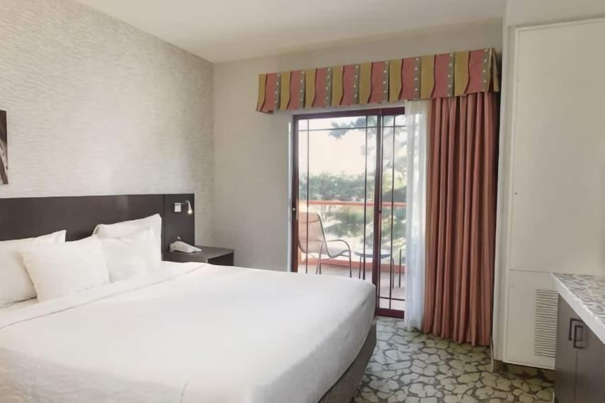 A hotel room with a large bed, a desk, a chair, a window with curtains, and a view of the outdoors.