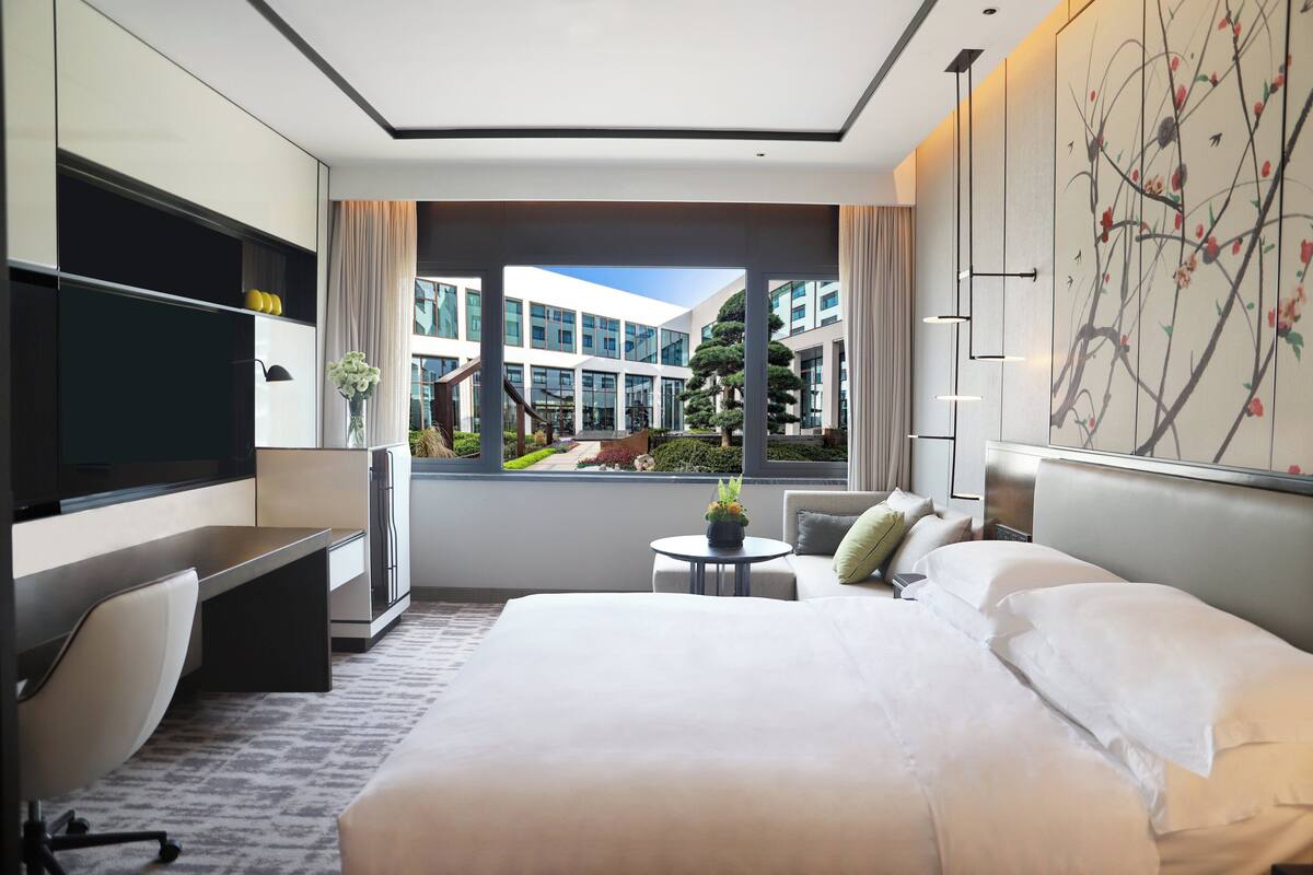 A modern hotel room with a large bed, a desk with a chair, a television, and a view of the building and greenery outside.