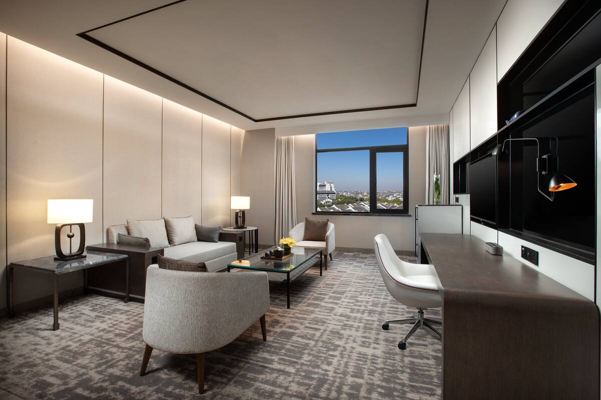 A modern hotel room with a large TV, comfortable seating, a glass coffee table, and a view of the city.