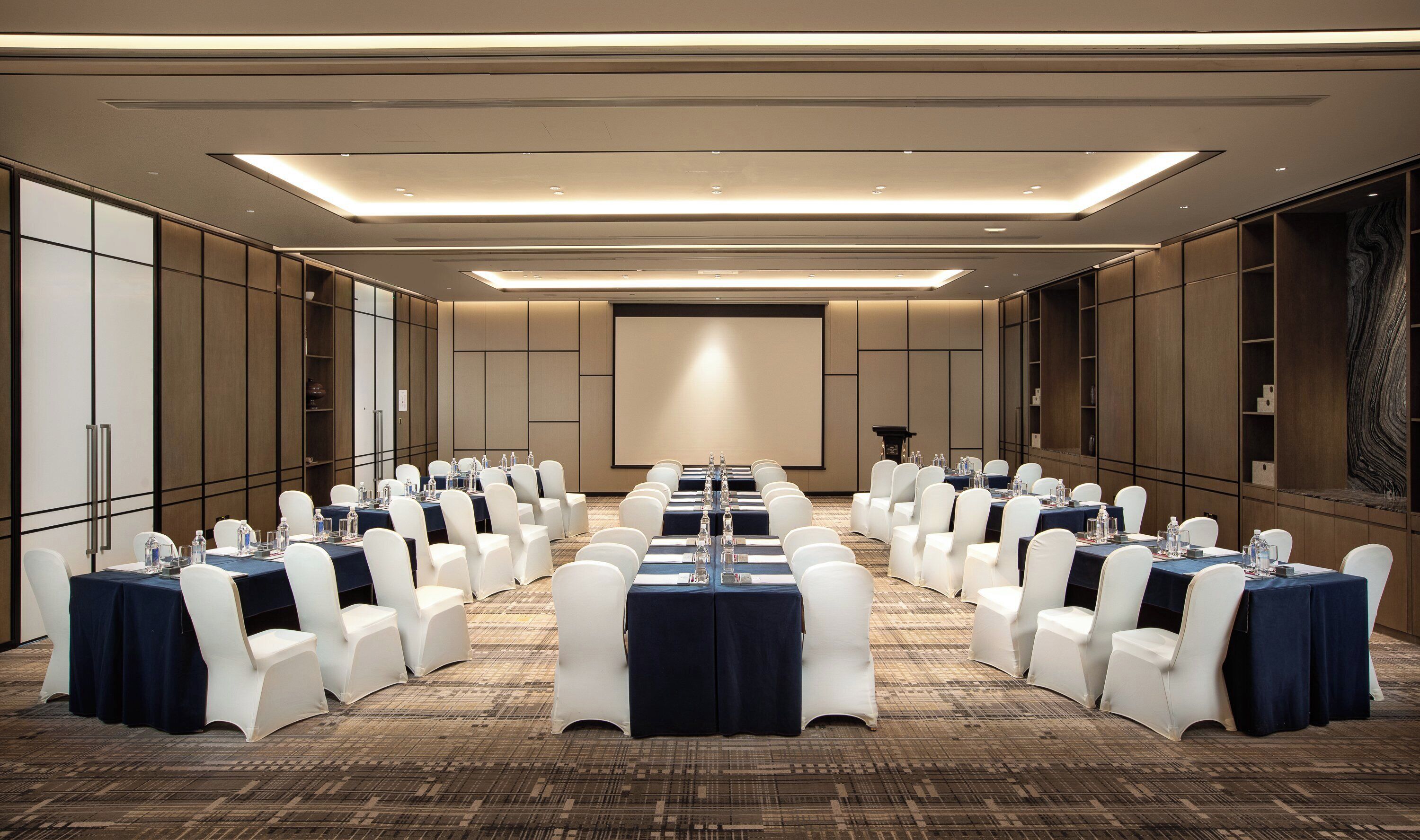 Un salón de conferencias con filas de mesas y sillas, una pantalla para proyector y un atril.