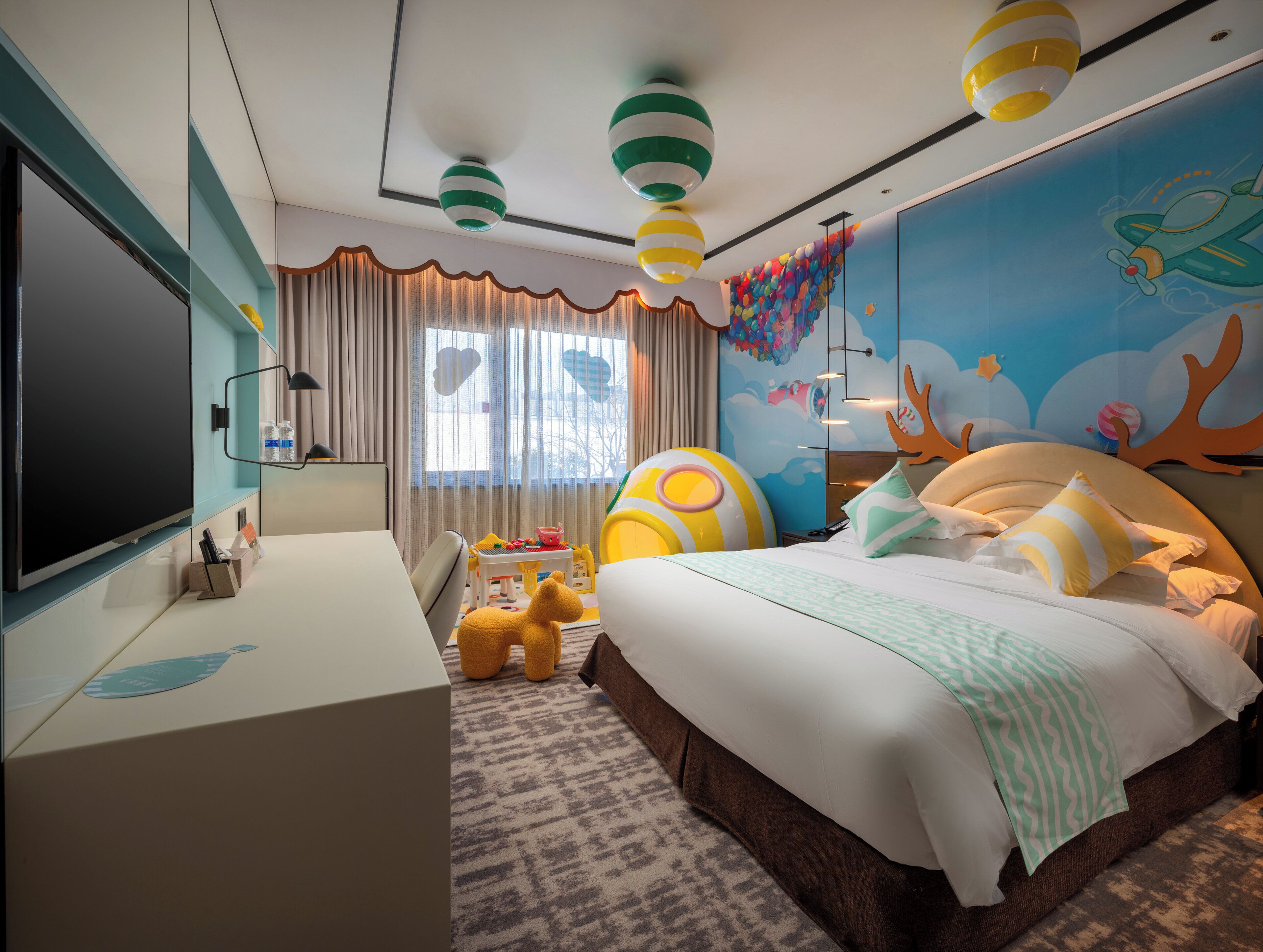 Una habitación de hotel con cama, escritorio, televisión, ventana con cortinas y un mural en la pared.