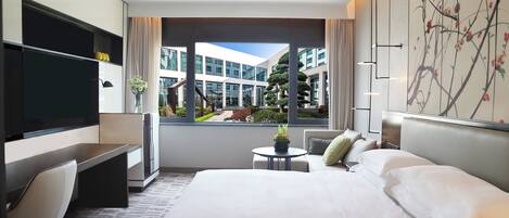 Habitación de hotel moderna con una cama grande, un escritorio con silla, televisor y vistas al exterior con edificios y vegetación.