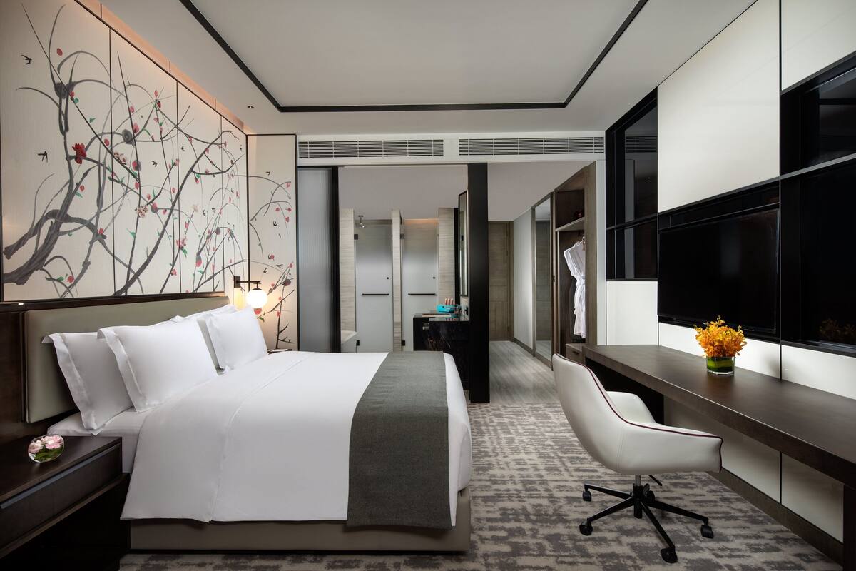 A modern hotel room with a large bed, a desk, a chair, a TV, and a decorative wall piece.