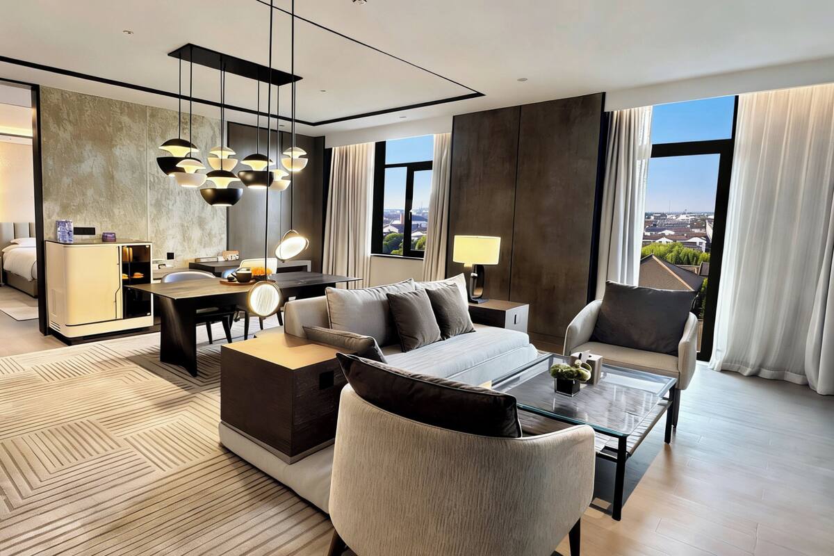 A modern hotel room with a sofa, armchair, and coffee table. There is a desk with a lamp and a view of the city through large windows.
