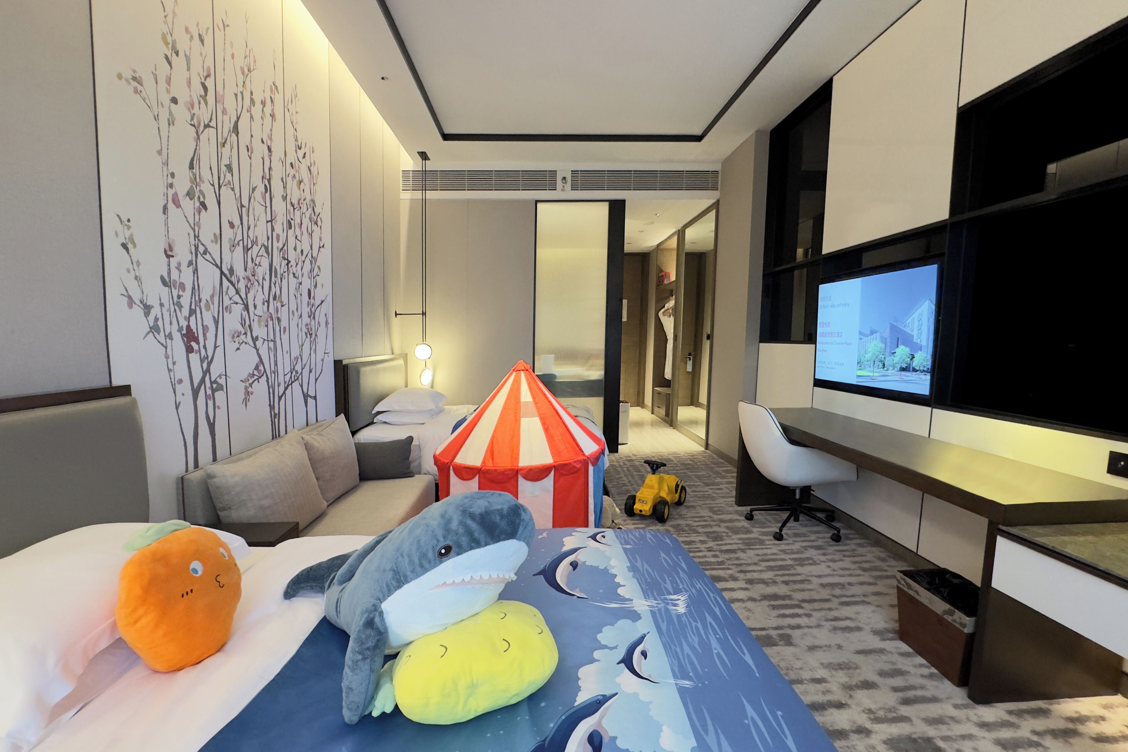 A hotel room with a bed, a desk, a TV, a small tent, and a toy car.