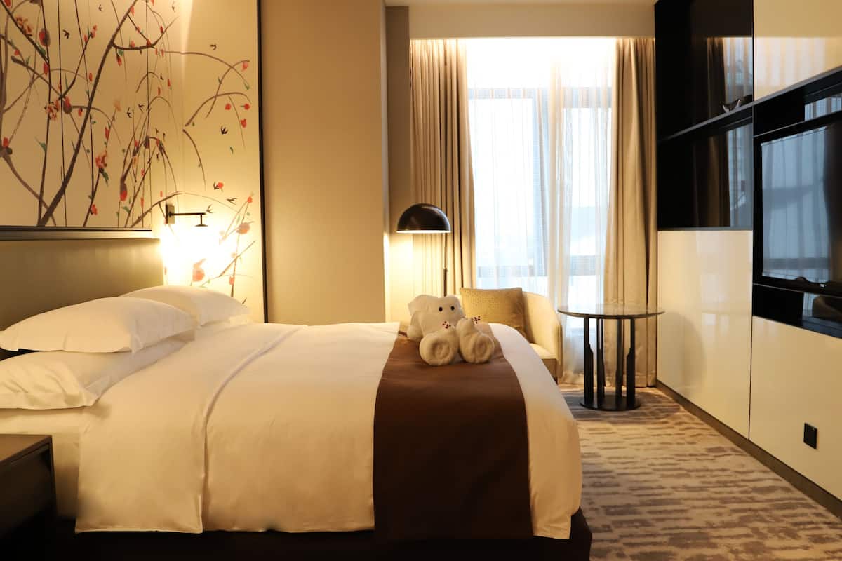 Ein Hotelzimmer mit einem ordentlich bezogenen Bett, einem Teddybären auf dem Bett, einem Fernseher an der Wand und einem Fenster mit Vorhängen.