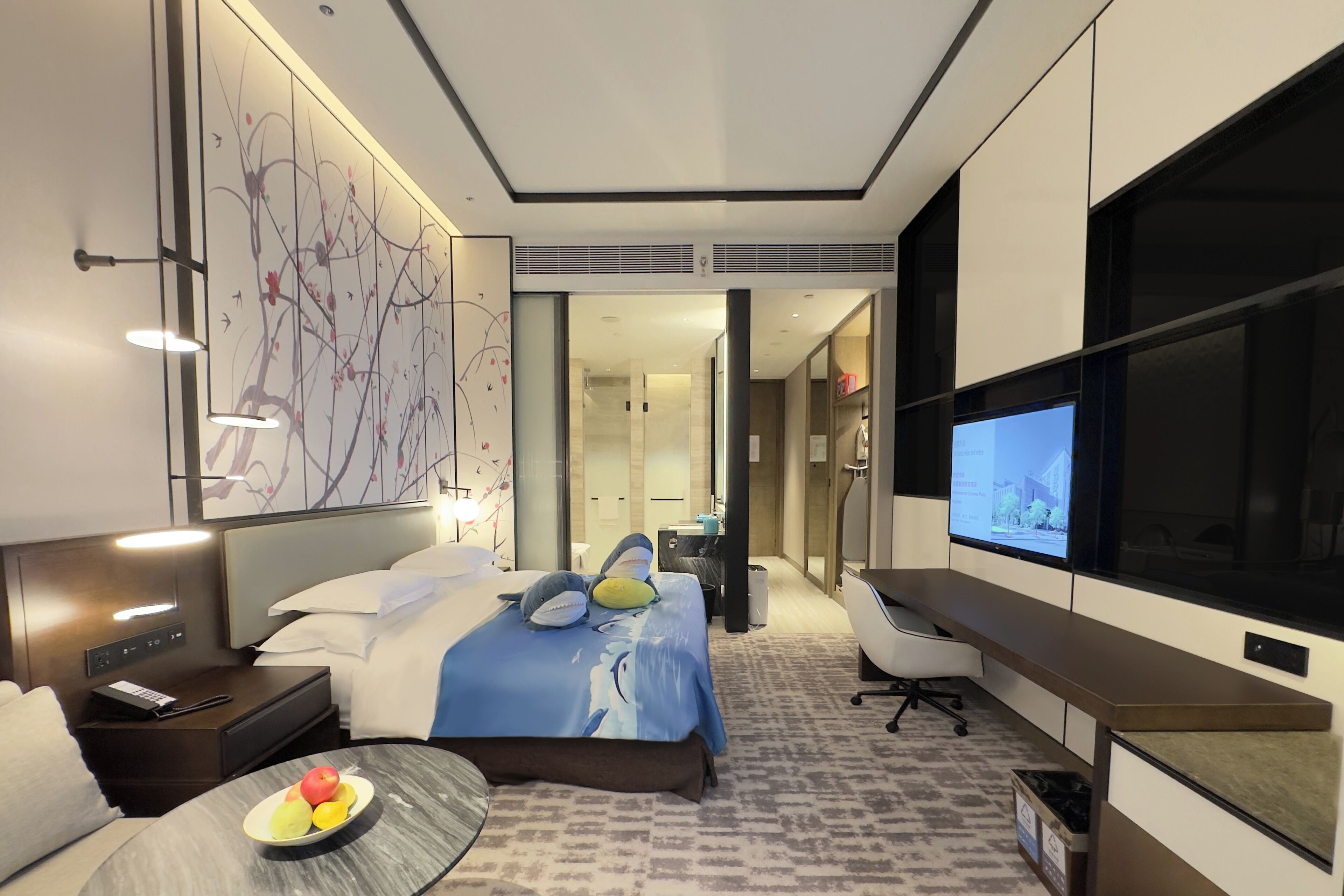 A modern hotel room with a bed, desk, TV, and a fruit bowl on the side table.