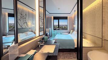 A modern hotel room with a large bed, a sitting area with a view, a bathroom with a bathtub, and a large window.