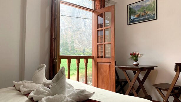 A room with a bed, a wooden door leading to a balcony, a small table with a vase, and a painting on the wall.