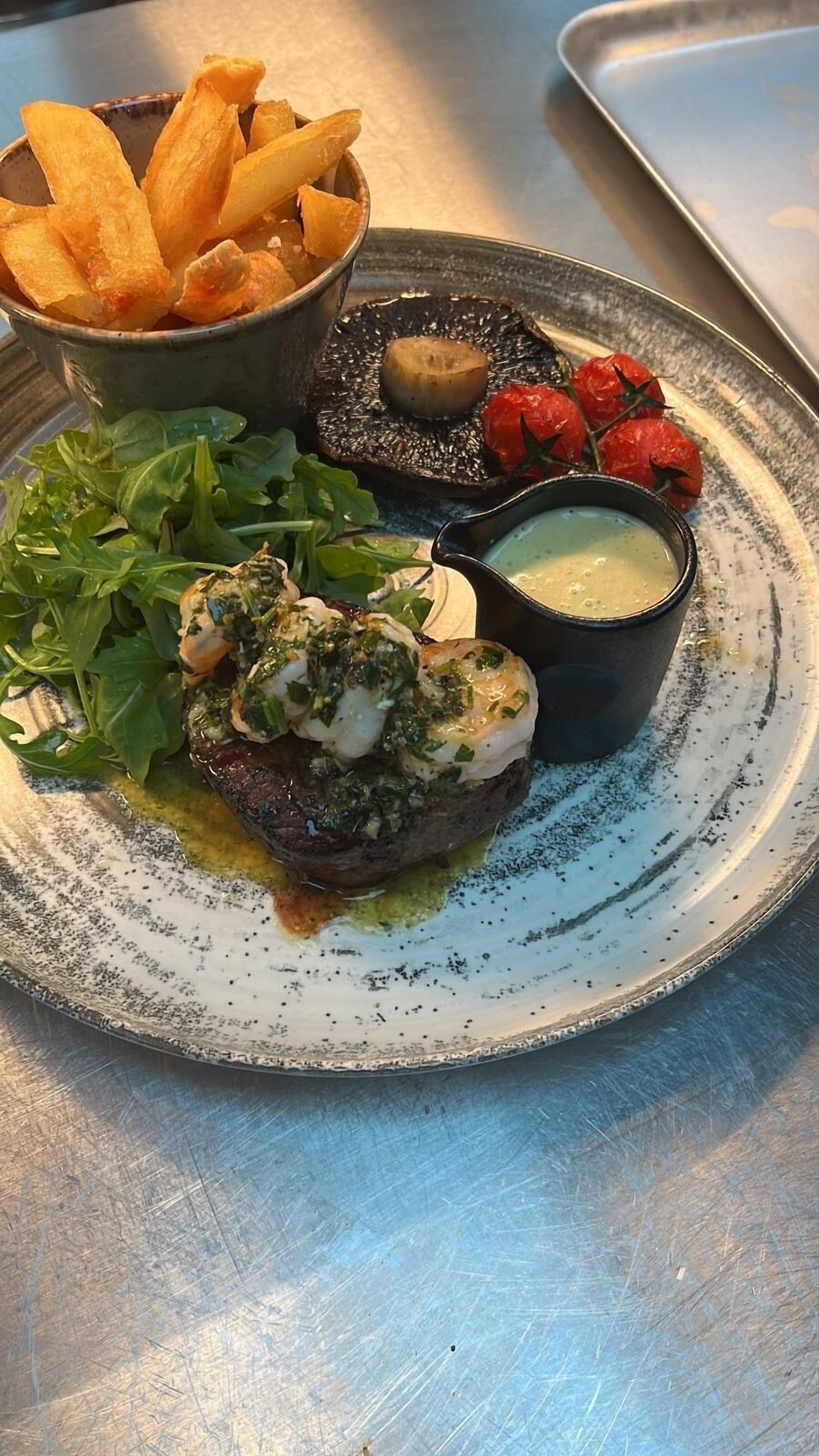 A plate with a steak topped with a green herb sauce, a bowl of fries, a grilled mushroom, and cherry tomatoes.