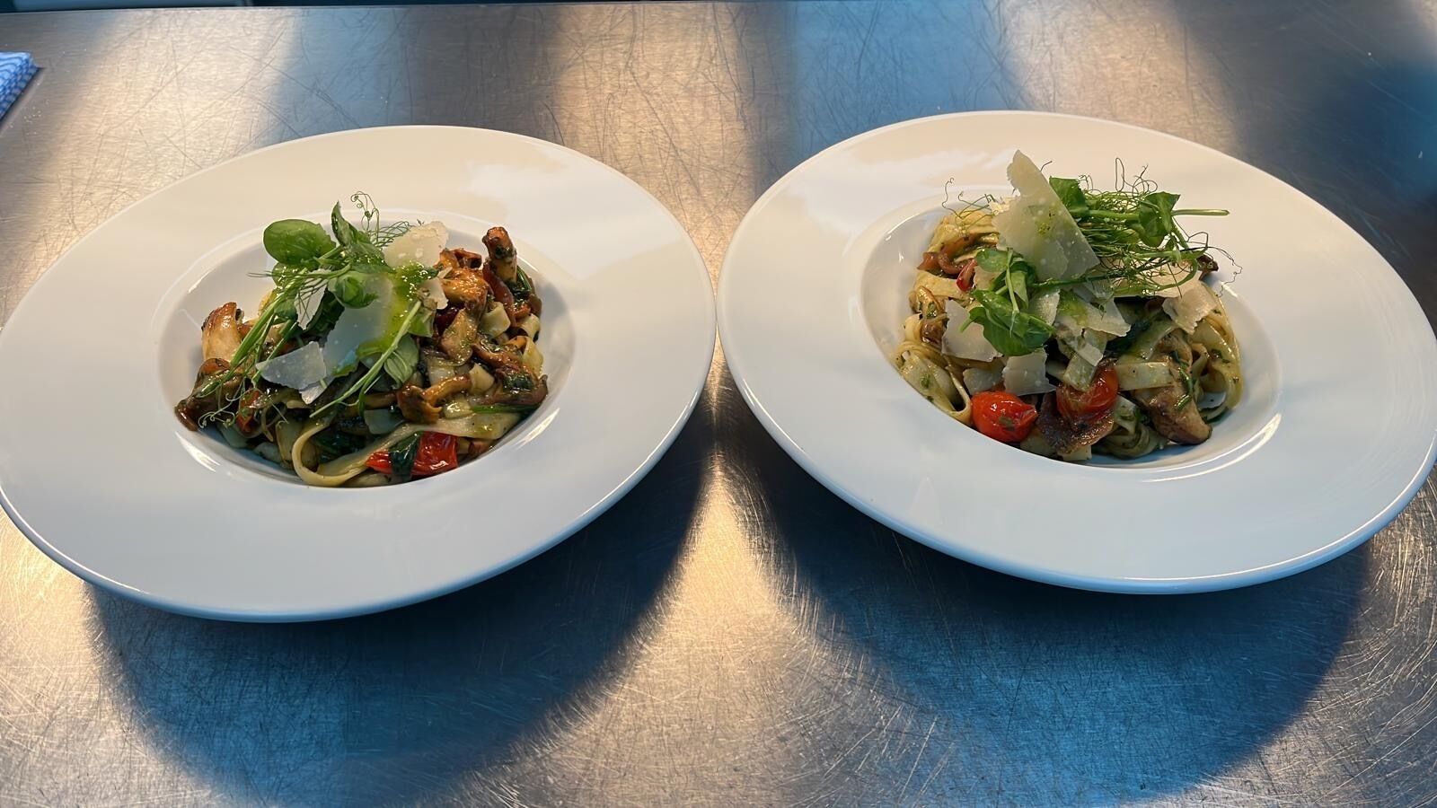 Two white plates of pasta with vegetables and greens on a metallic surface.
