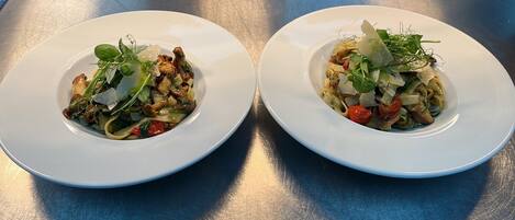 Two white plates of pasta with vegetables and greens on a metallic surface.