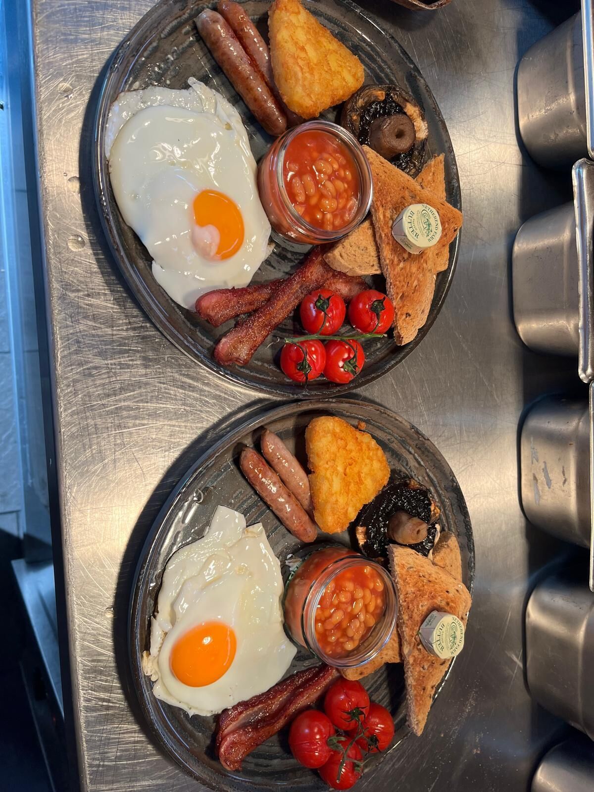 Two plates of breakfast food including eggs, sausages, bacon, mushrooms, toast, and tomatoes.
