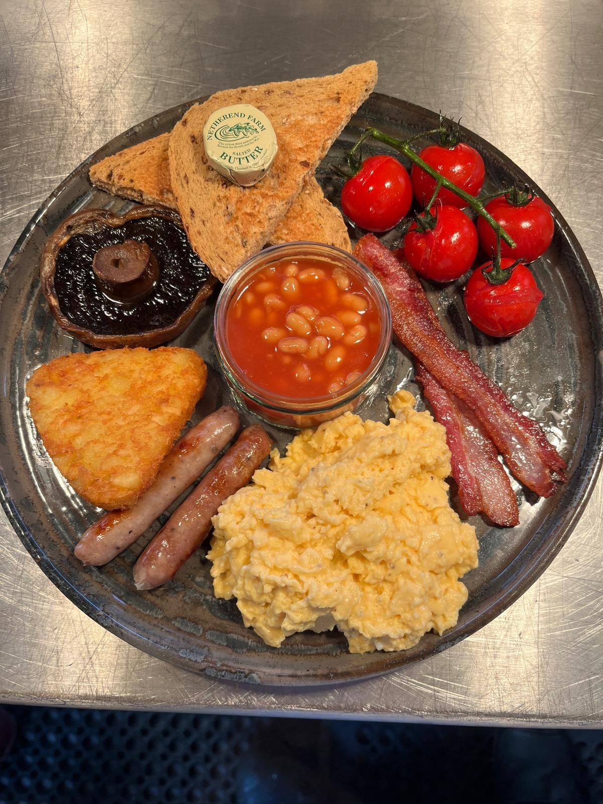 Een bord met diverse ontbijtartikelen, waaronder toast, bonen, spek, worstjes, roerei en tomaten.