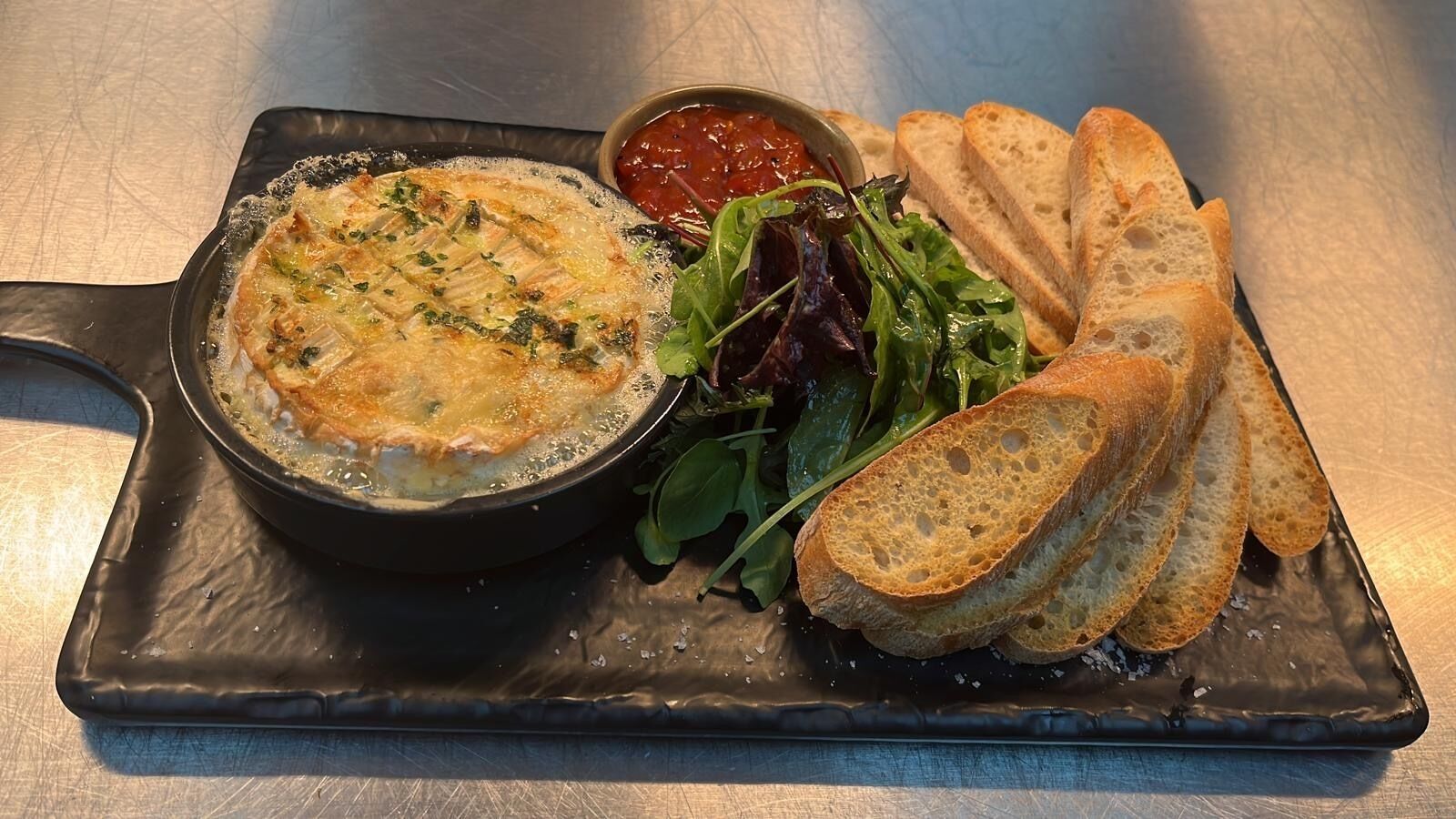 A dish of baked fish with a side of greens and bread on a black plate.