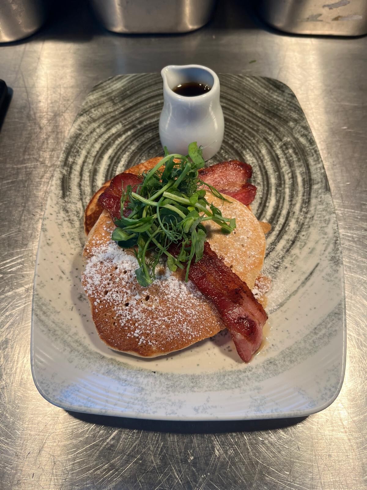 A plate with pancakes topped with bacon, greens, and a small bowl of syrup.