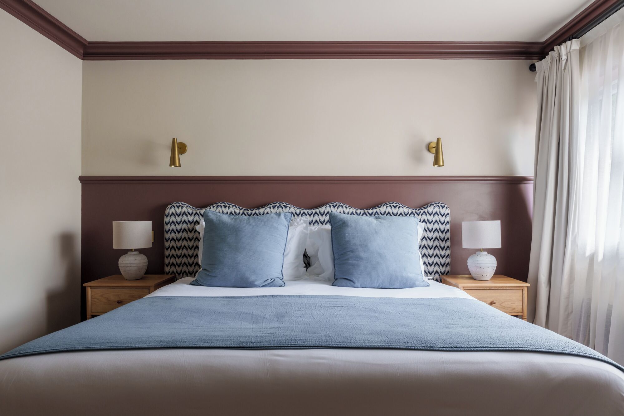 Una cama bien hecha con un edredón azul y dos almohadas, flanqueada por mesitas de noche con lámparas, en una habitación de diseño sencillo y limpio.