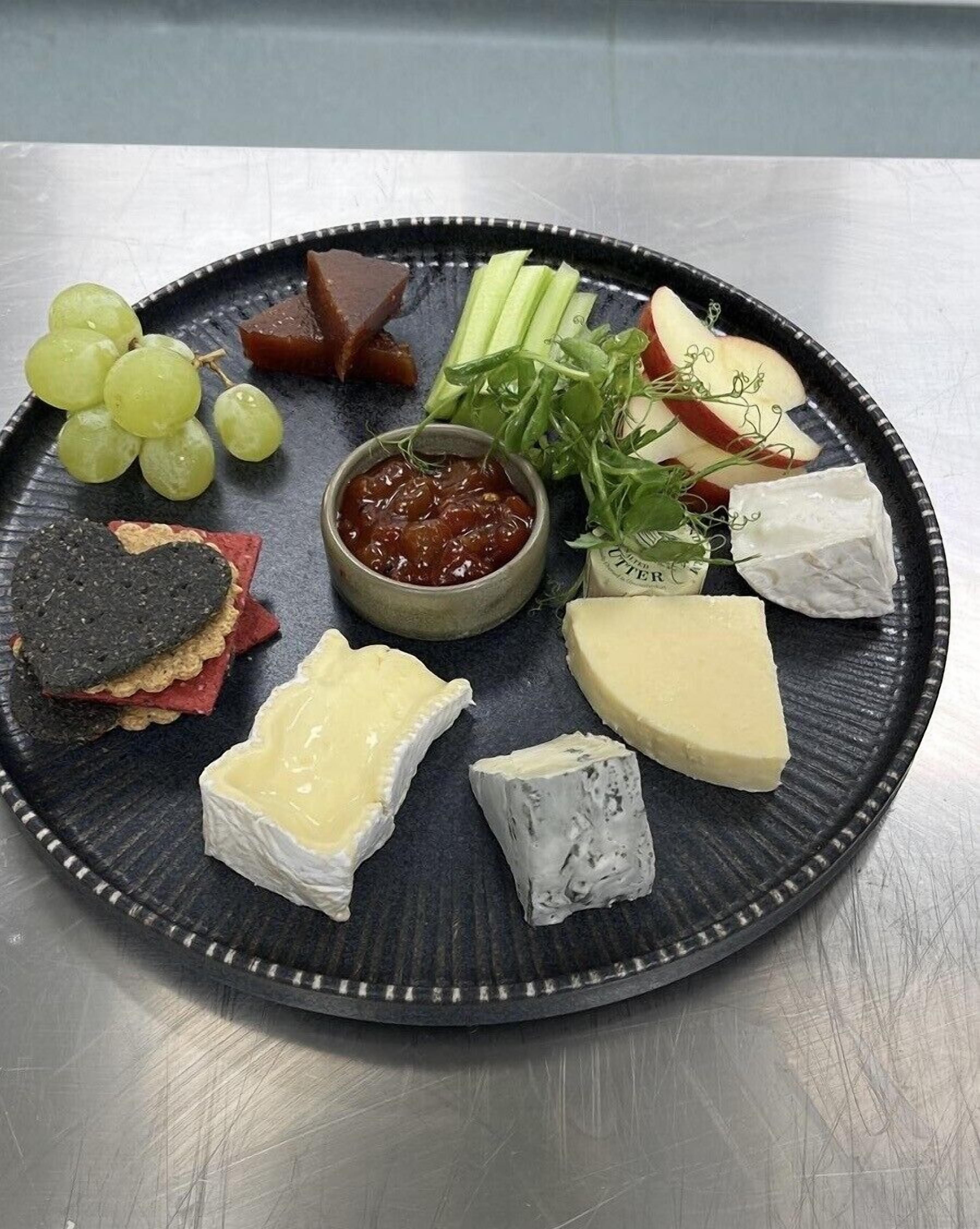 Una tabla de embutidos con varios quesos, uvas, manzanas y un recipiente pequeño de chutney.