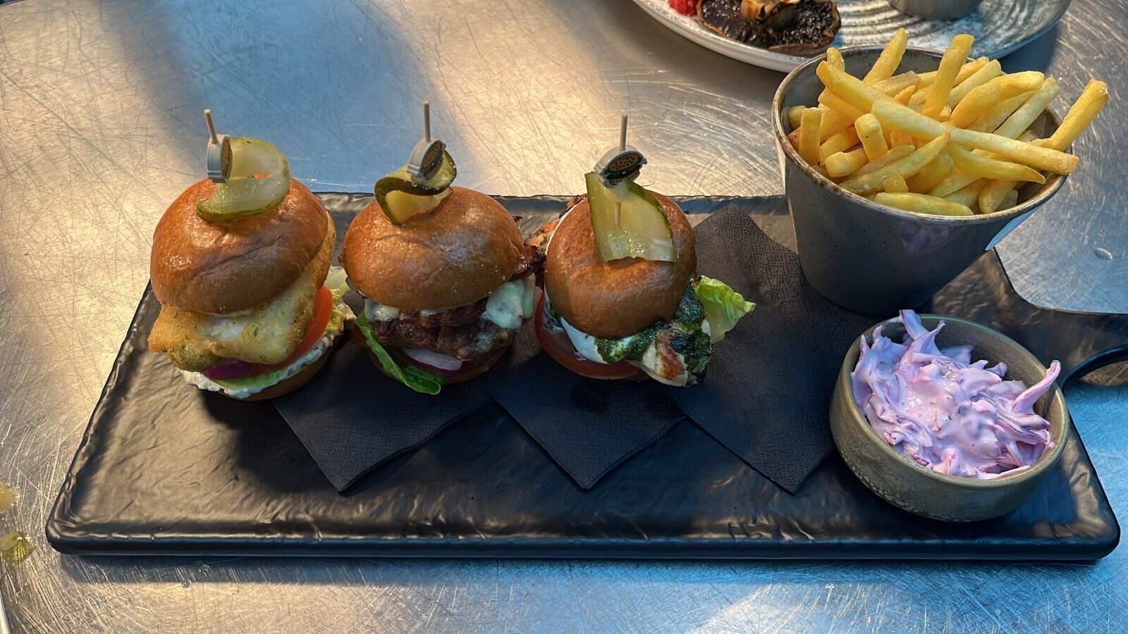 Three burgers with pickles on top, a bowl of fries, and a small bowl of coleslaw on a metal tray.