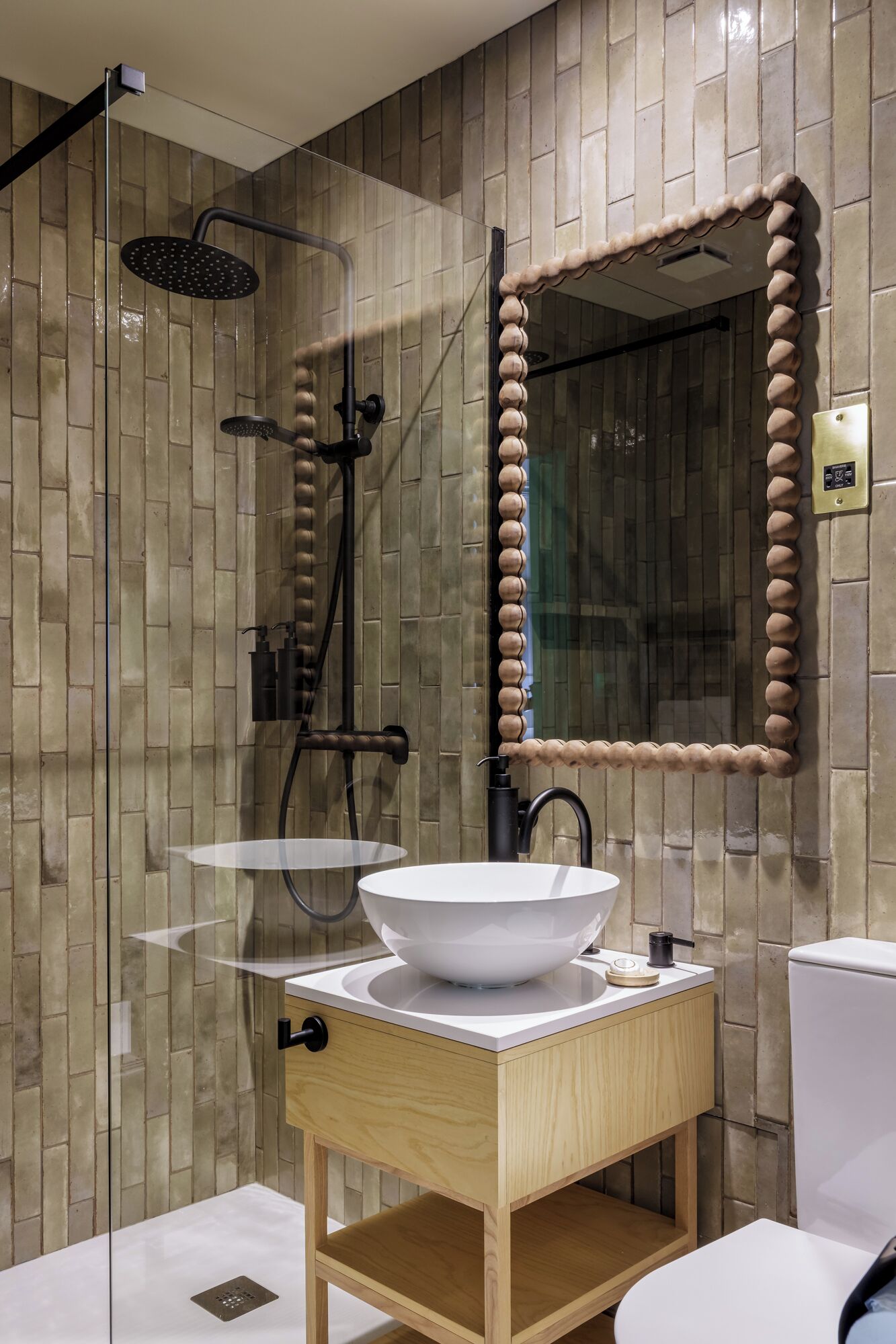 Un baño moderno con ducha envuelta en vidrio, lavamanos blanco con tocador de madera y un espejo grande con marco decorativo.