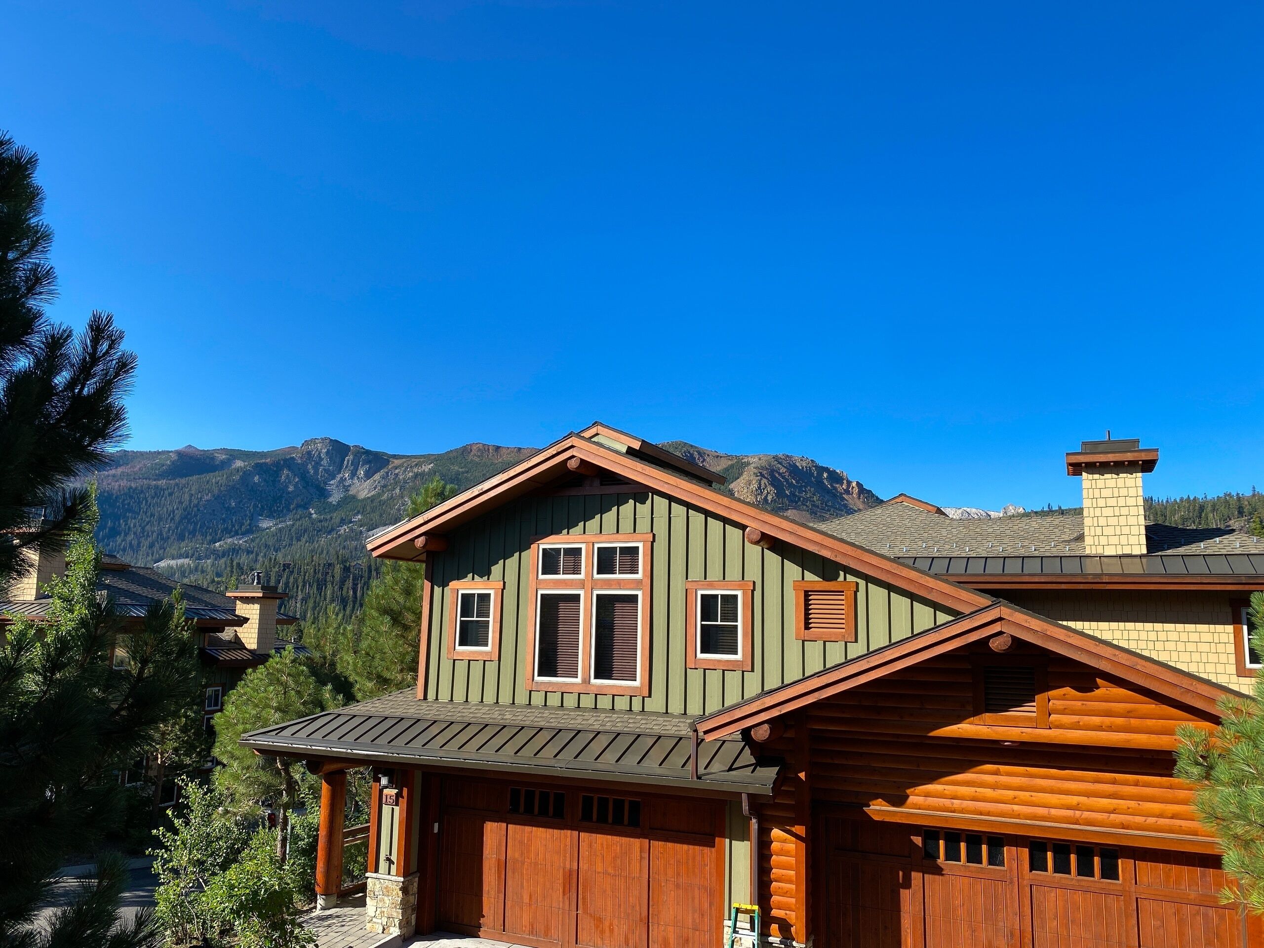 A two-story log cabin with a green exterior and brown trim, featuring a large garage and a mountain backdrop.