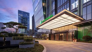 A modern hotel entrance with a glass facade, a covered entrance area, and a landscaped garden with neatly trimmed bushes and a tree. - Holiday Inn Hangzhou Binjiang (Hangzhou)